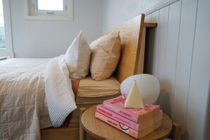 A bedroom with a bed, pillows, a bedside table with books, and a window. - Carinya Luxe Cottage (Newstead)
