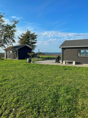 A grassy area with two small buildings, one with a window and another with a door, and a bench. - Holiday cottage with wild bath by field and water (Kerteminde)