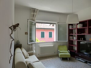 A living room with a sofa, a green armchair, a television, a bookshelf, and a window with a view of a pink building. - Beautiful Apartment in Jesolo, PVT Parking (Jesolo)