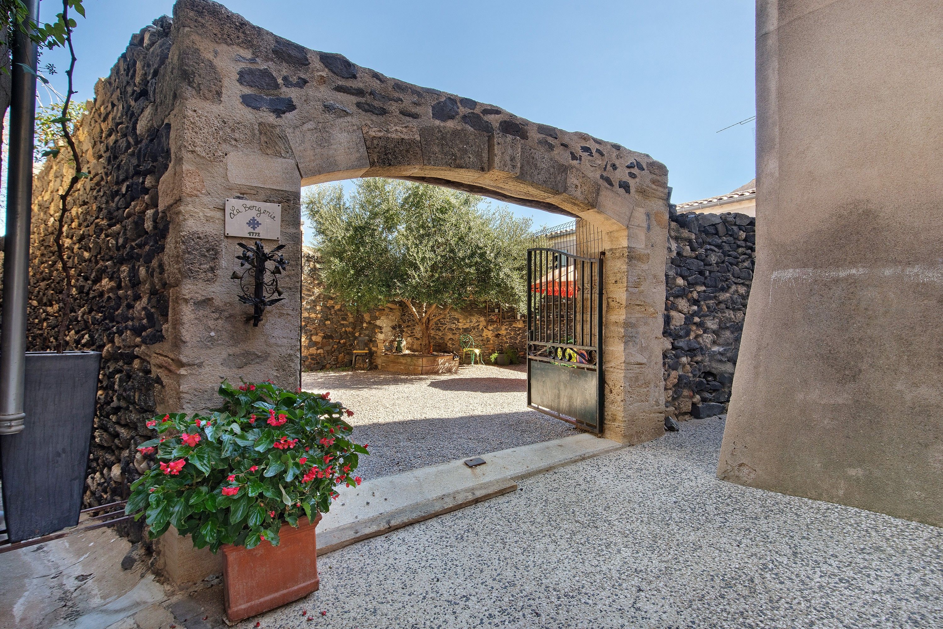 A stone archway leading to a courtyard with a gravel path, a potted plant with red flowers, and a sign indicating the year 1772.