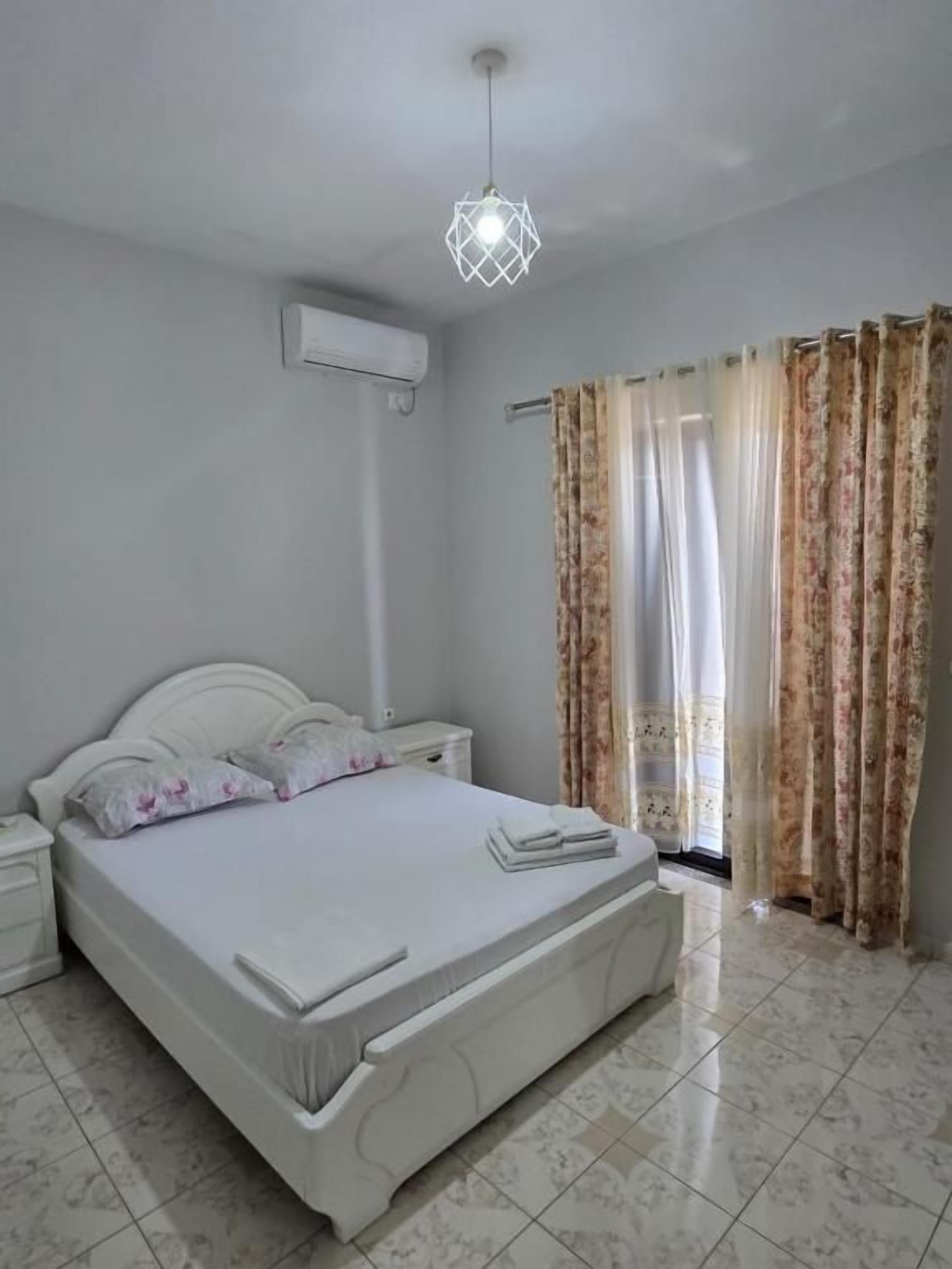 A bedroom with a bed, nightstands, a hanging light fixture, an air conditioning unit, and a window with curtains.
