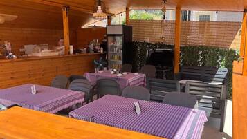 Un área de comedor al aire libre con mesas y sillas de madera, una parrilla y una barra con una persona detrás del mostrador.