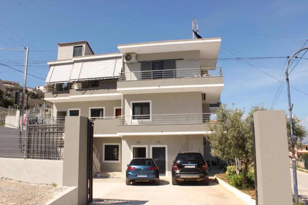 A two-story residential building with a balcony on the upper floor, a gated entrance, and two parked cars.