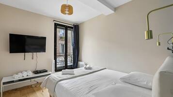 Ein modernes Schlafzimmer mit einem Bett, einem Flachbildfernseher, einer Bank mit Handtüchern und einem Fenster mit Blick auf ein Gebäude.