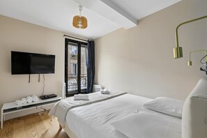 A modern bedroom with a bed, a flat-screen TV, a bench with towels, and a window with a view of a building. - Charming Apartment with Cozy Open-Plan Living (Boulogne-Billancourt)