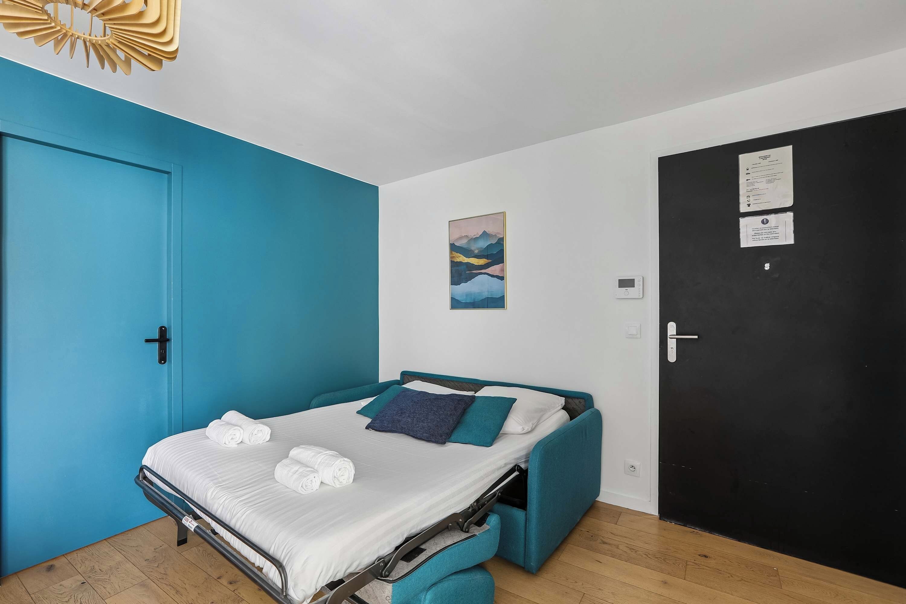 A compact bedroom with a bed, a blue wall, a black door, and a small painting.