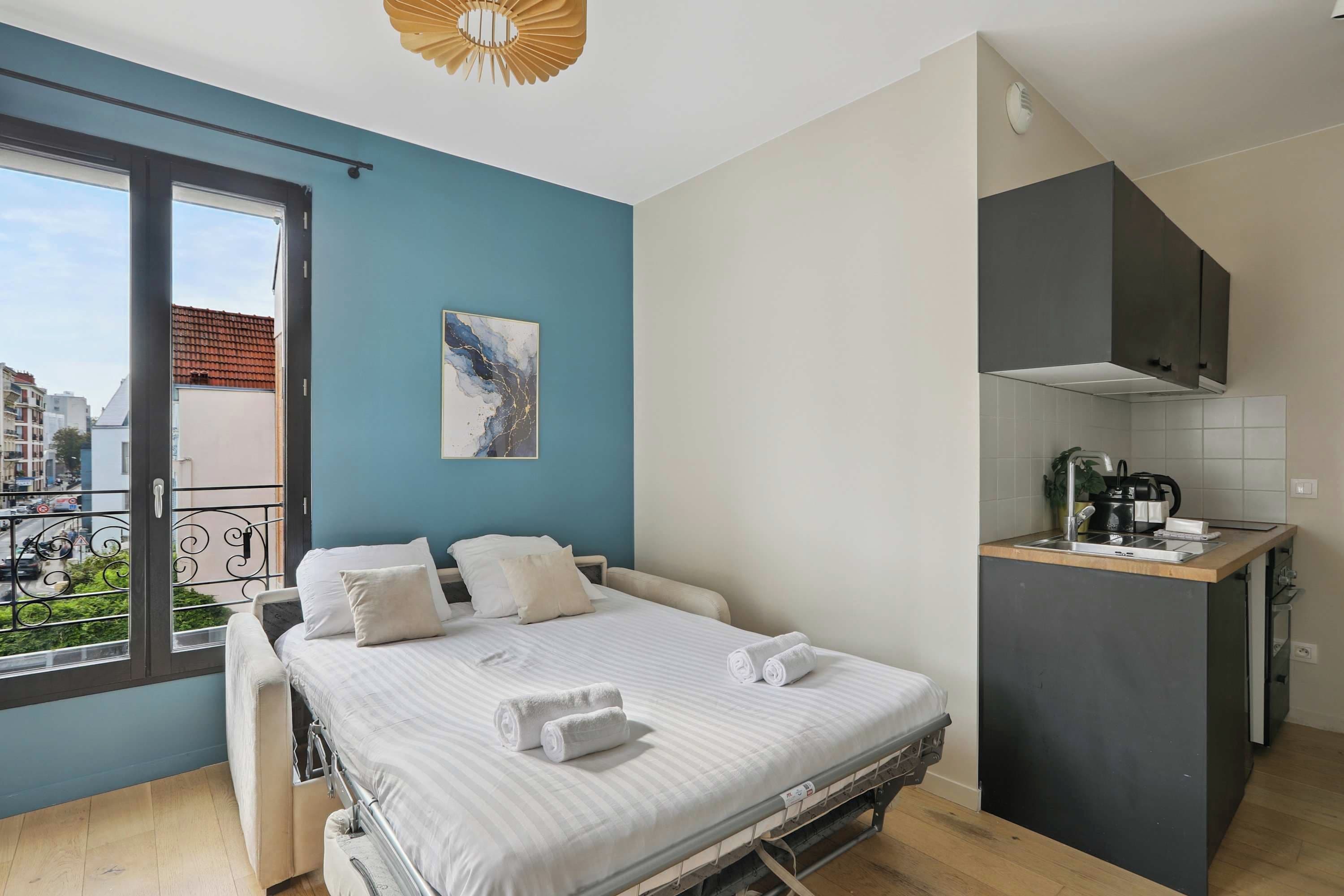 Une chambre avec un lit, une petite kitchenette, une fenêtre donnant sur des immeubles et un luminaire au plafond.