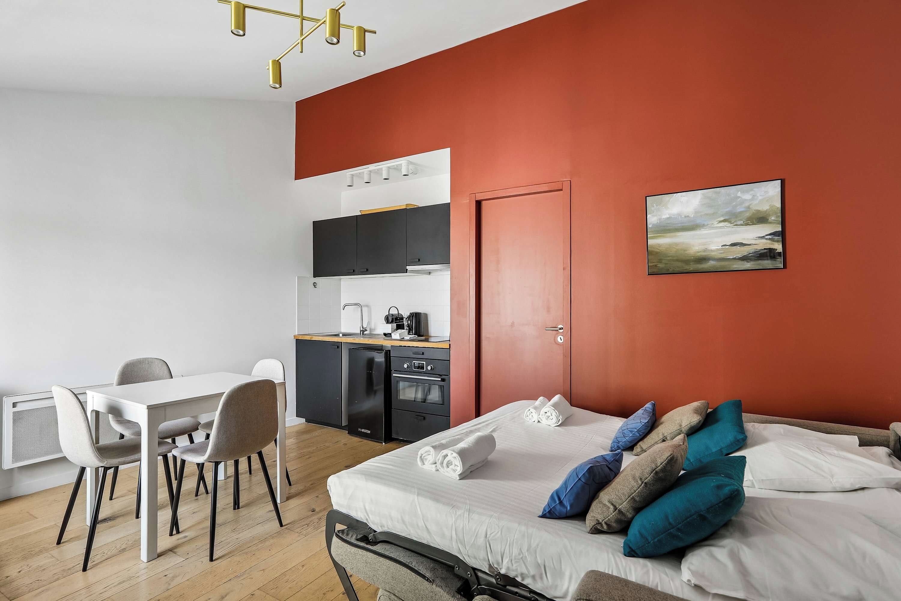 A modern bedroom with a bed, pillows, a desk, and a kitchenette.
