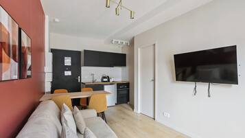 A modern living room with a sofa, a dining table with chairs, a kitchen area with cabinets and appliances, and a mounted television.