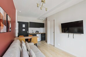 A modern living room with a sofa, a dining table with chairs, a kitchen area with cabinets and appliances, and a mounted television. - Stylish Apartment with Cozy Bedroom & Sofa Bed (Boulogne-Billancourt)