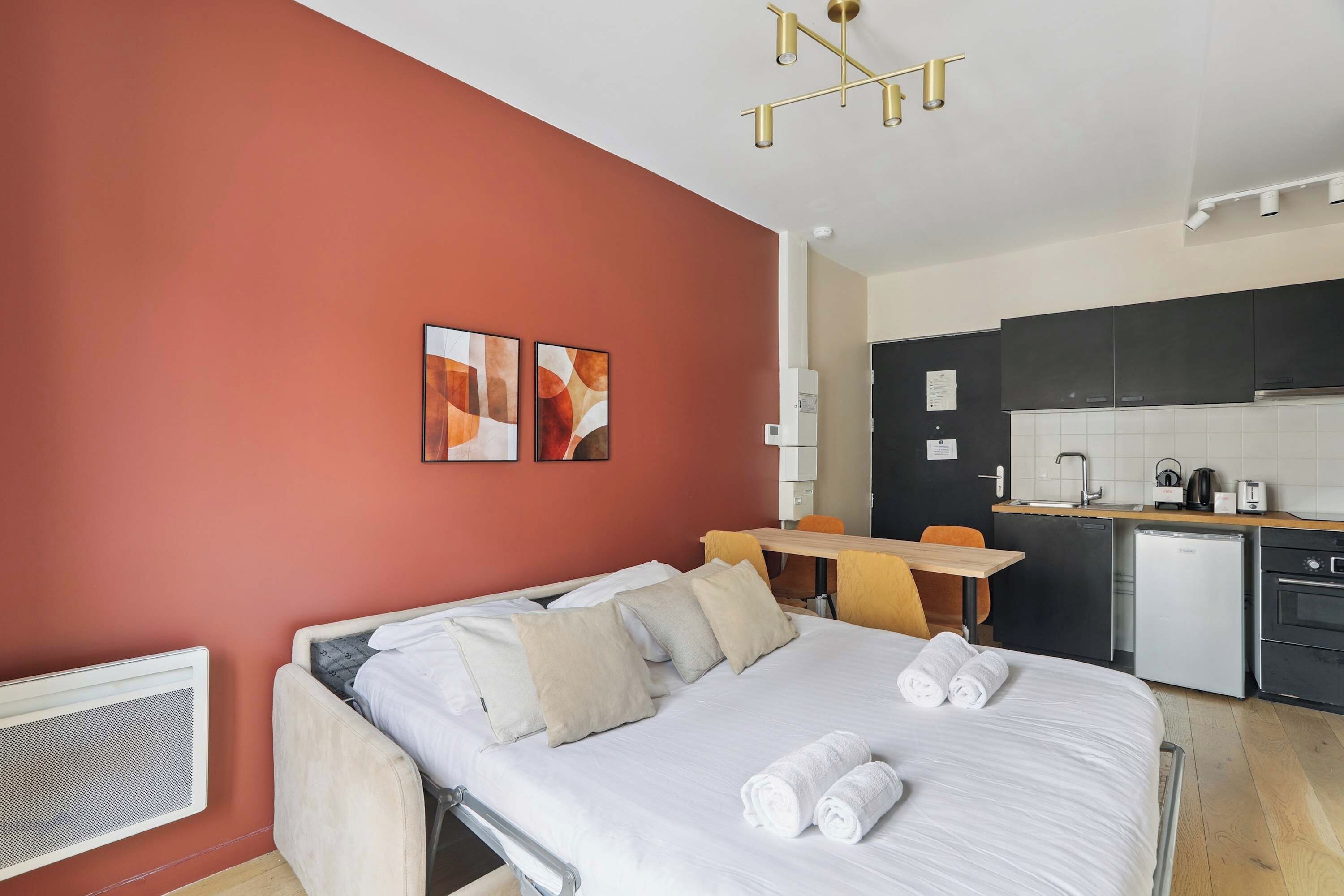 A hotel room with a bed, a desk, a chair, a kitchenette, and artwork on the wall.