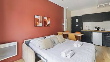 A hotel room with a bed, a desk, a chair, a kitchenette, and artwork on the wall.