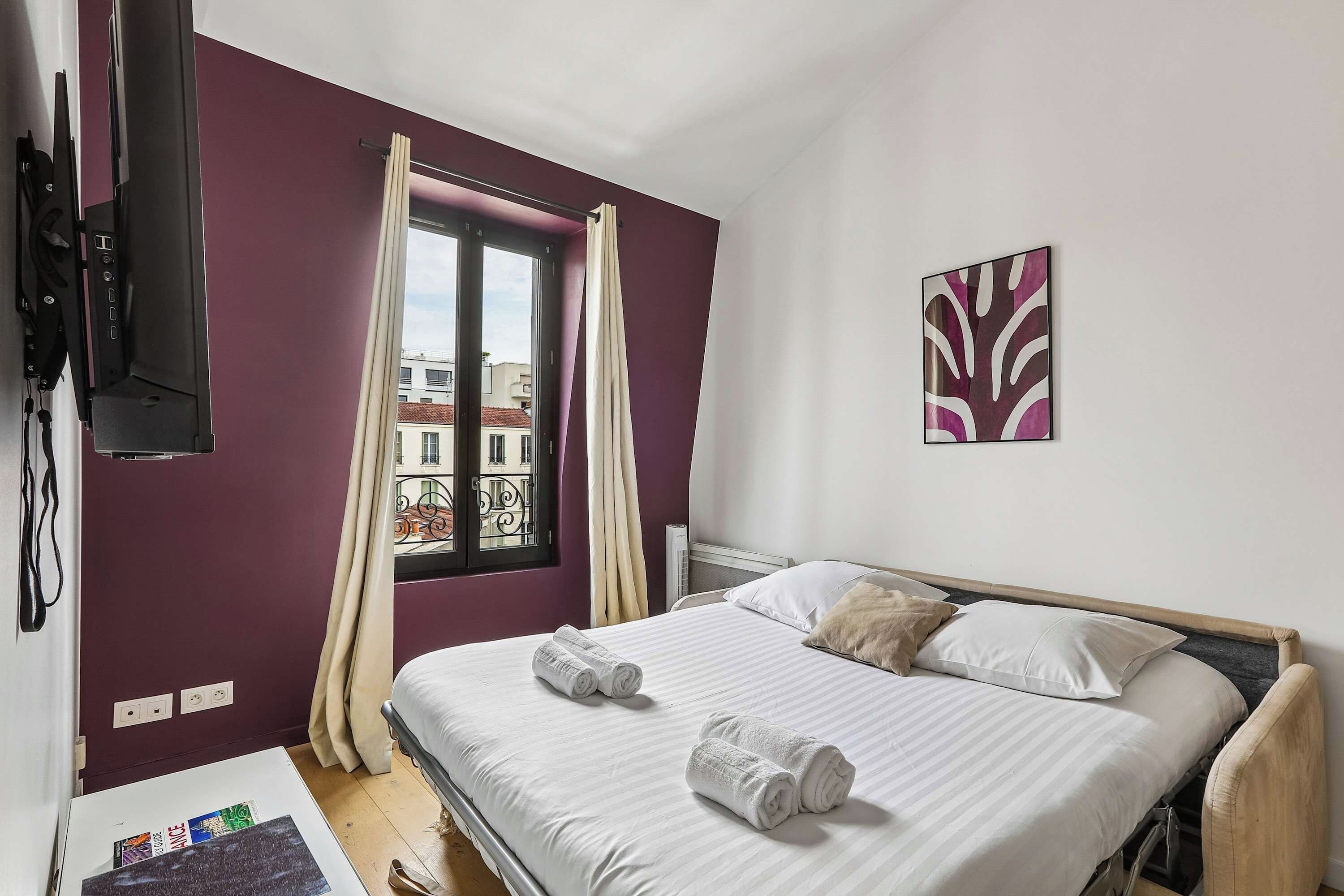 A hotel room with a bed, a window with curtains, a mounted TV, and a wall-mounted artwork.
