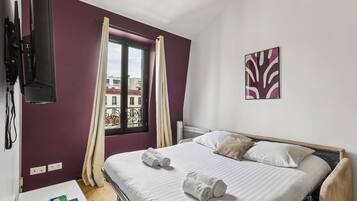 A hotel room with a bed, a window with curtains, a mounted TV, and a wall-mounted artwork.