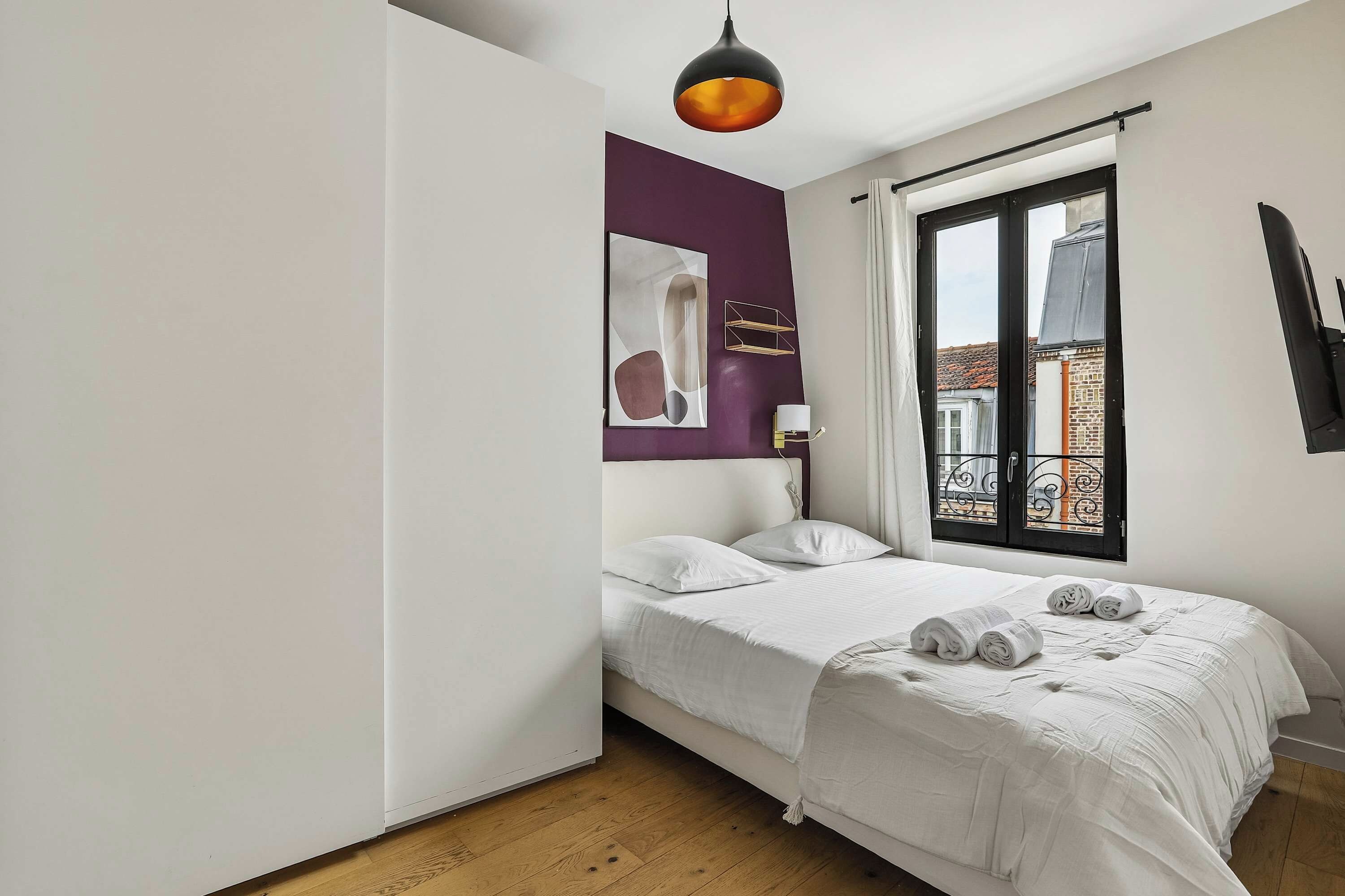 A bedroom with a bed, a hanging light fixture, a window with a view of buildings, and a wall-mounted shelf.