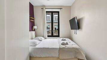 Ein Schlafzimmer mit einem Bett, einem an der Wand montierten Fernseher und einem Fenster mit Blick auf Gebäude.