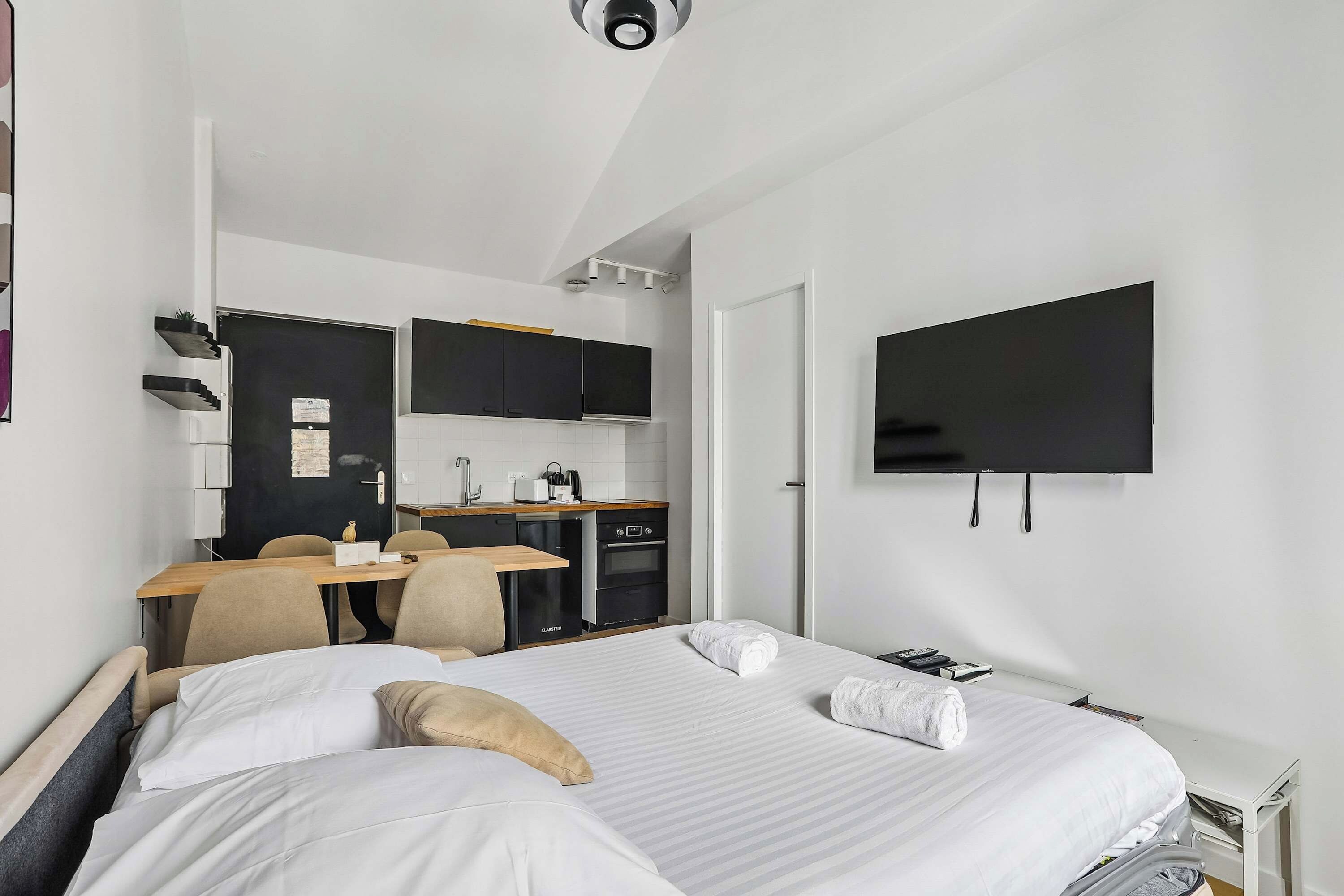 A hotel room with a bed, a desk with a chair, a kitchenette, and a flat-screen TV mounted on the wall.
