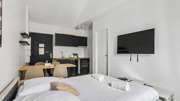 A hotel room with a bed, a desk with a chair, a kitchenette, and a flat-screen TV mounted on the wall.