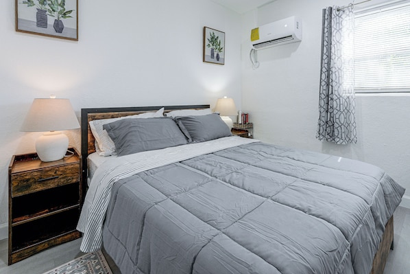 A neatly made bed with a grey quilt, a bedside lamp, and a nightstand with a book and a small lamp.