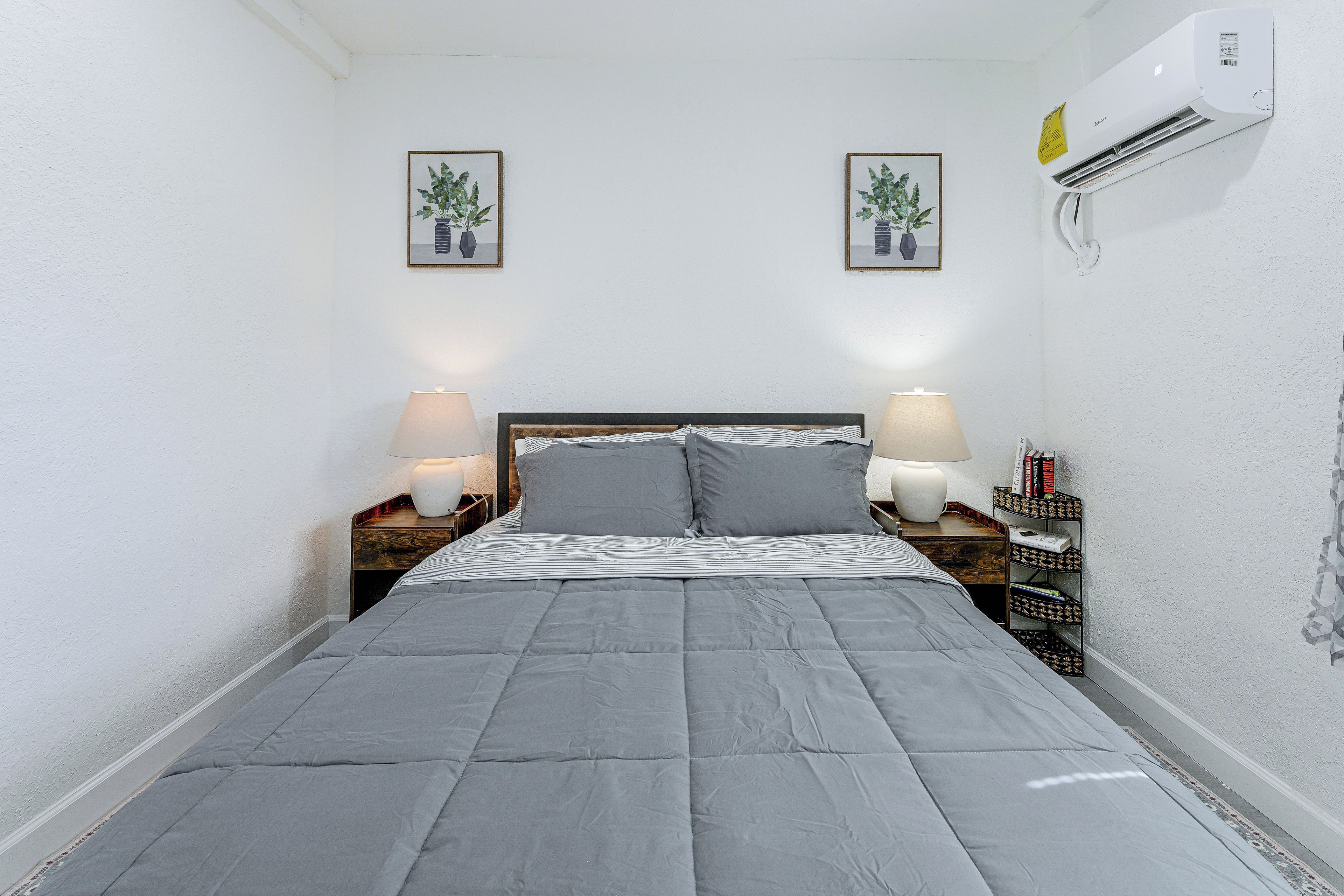 A bedroom with a bed, two bedside lamps, a wall-mounted air conditioner, and two framed pictures.
