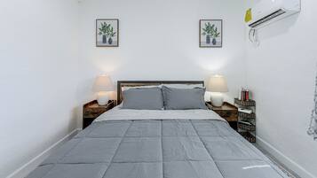 A bedroom with a bed, two bedside lamps, a wall-mounted air conditioner, and two framed pictures.
