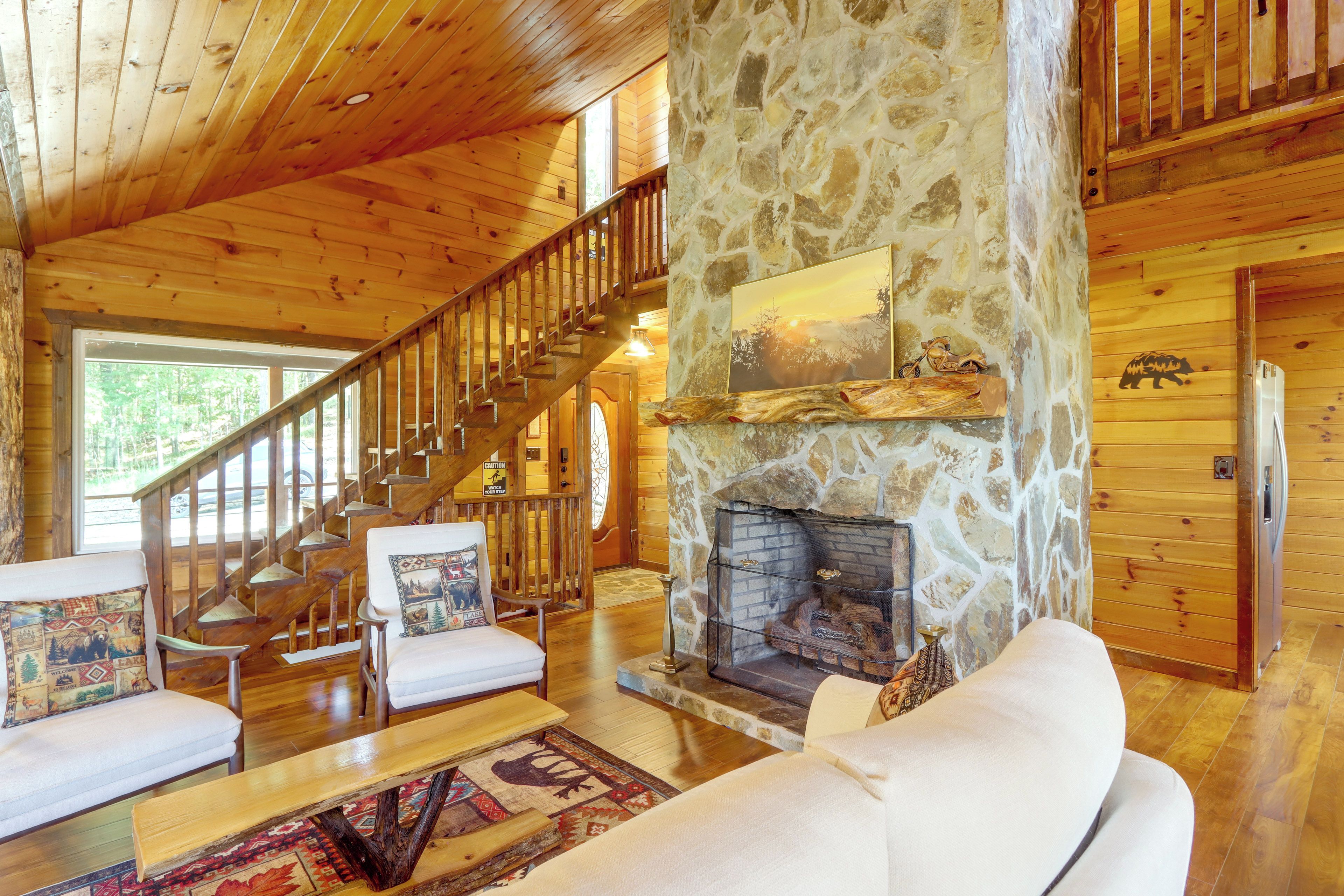 A log cabin interior with a stone fireplace, wooden staircase, and a cozy seating area.