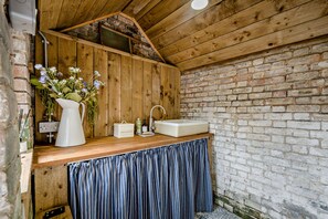 A rustic bathroom with a wooden ceiling, a brick wall, a wooden vanity with a sink, and a vase of flowers. - Host Stay Silk Weaver s Cottage (Norwich)