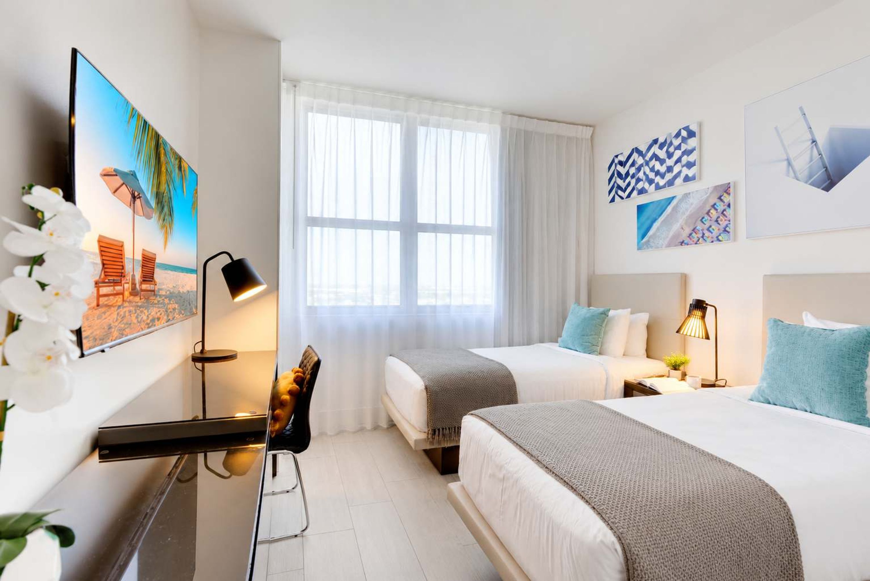 A hotel room with two beds, a desk, a chair, a window with sheer curtains, and artwork on the walls.
