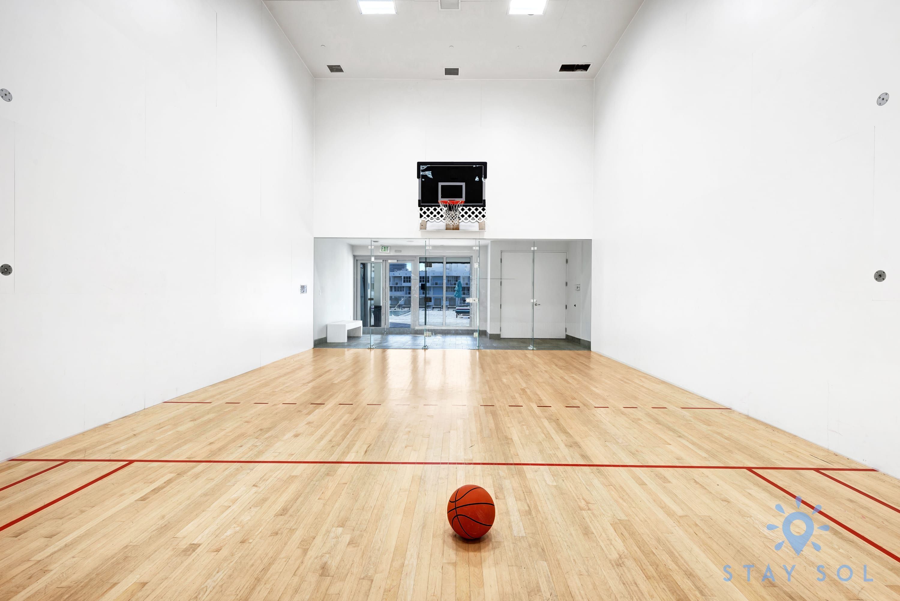 A spacious indoor basketball court with a wooden floor, a basketball hoop, and a clear view of the outside through glass doors.