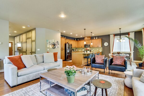 A modern living room with a sofa, coffee table, and chairs, a kitchen with wooden cabinets and stainless steel appliances, and a dining area with a table and chairs.