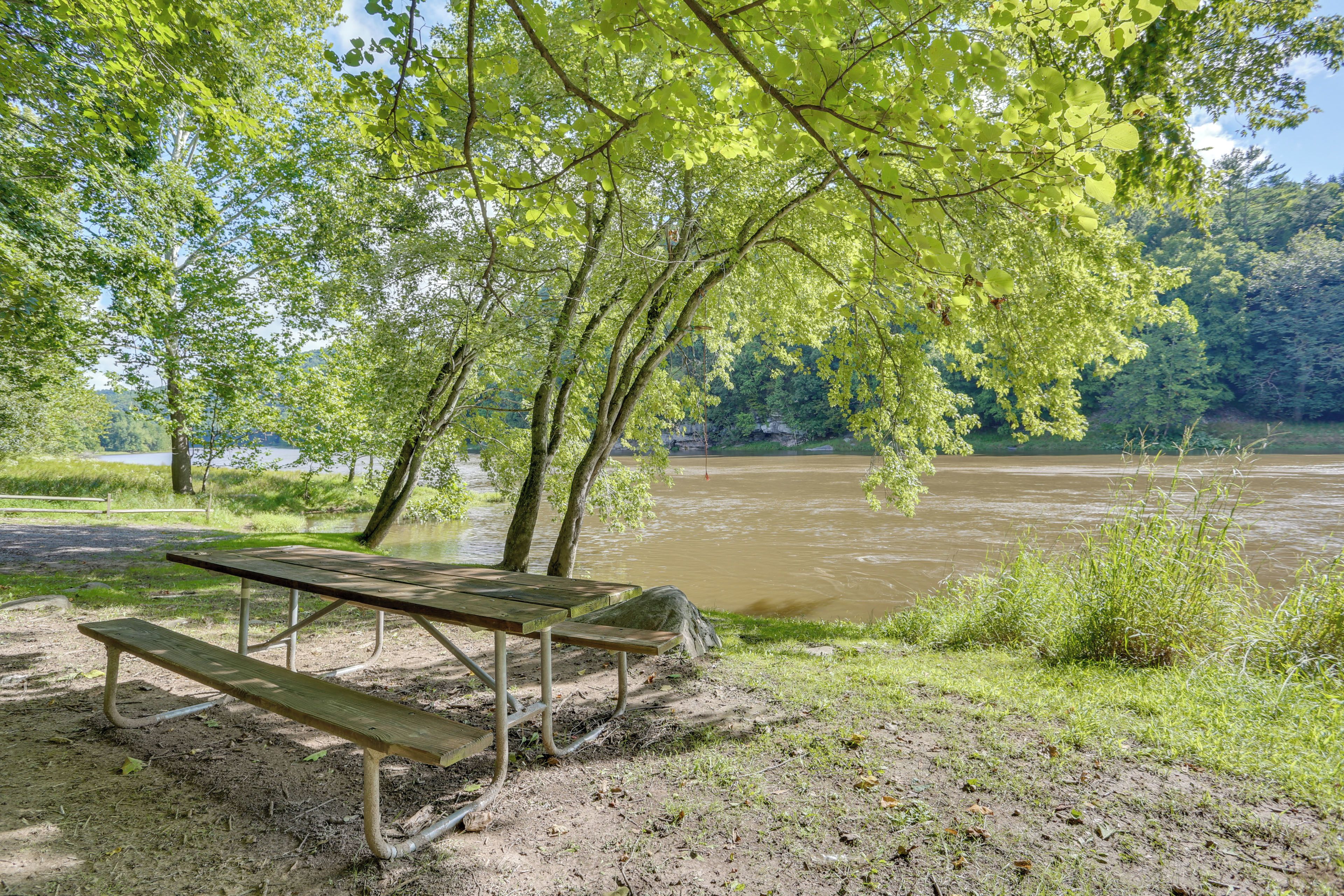 A picnic table with benches by a river, surrounded by trees and a grassy area.