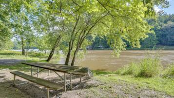 A picnic table with benches by a river, surrounded by trees and a grassy area.