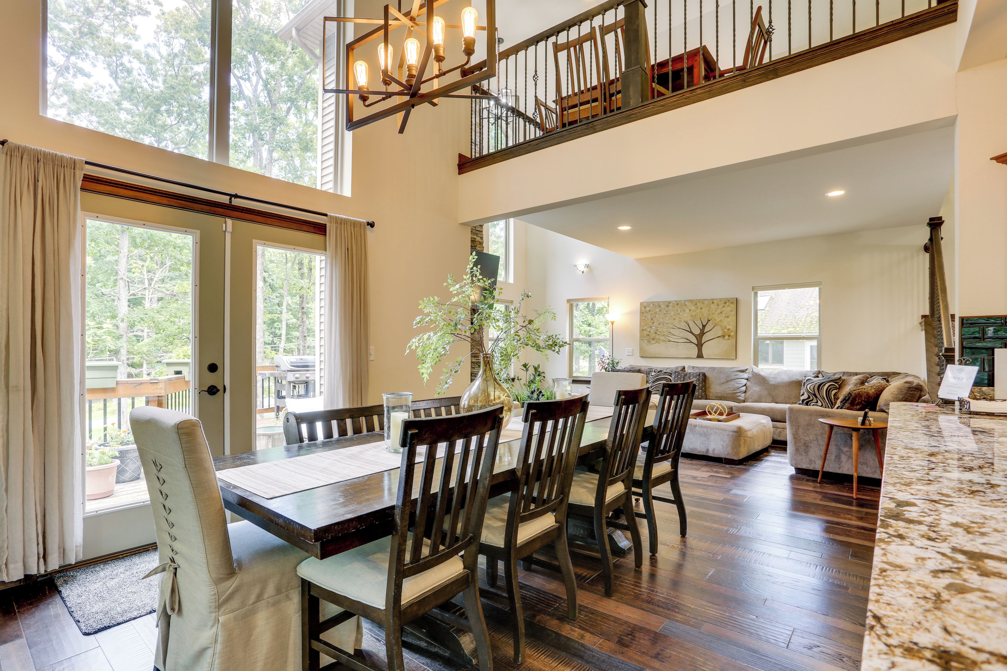 A spacious dining area with a high ceiling, wooden dining table, and chairs. A large window provides a view of the outdoors.
