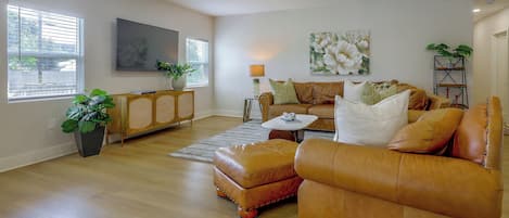 Un salón espacioso con un ventanal grande, un televisor de pantalla plana, un conjunto de sofás, una mesa de centro y un cuadro en la pared.