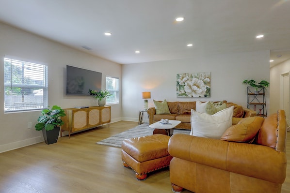 A spacious living room with a large window, a flat-screen TV, a sofa set, a coffee table, and a painting on the wall.