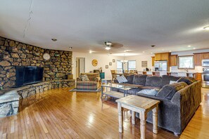 Un salón amplio con pared de piedra, televisor de pantalla plana, sofá grande y una zona de comedor con armarios de madera y una cocina al fondo.