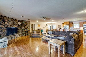 Un salón amplio con pared de piedra, televisor de pantalla plana, sofá grande y una zona de comedor con armarios de madera y una cocina al fondo.