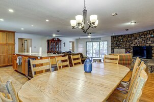 A spacious dining area with a wooden table and chairs, a chandelier, and a stone wall with a television.