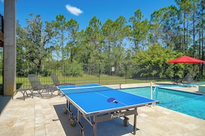 Mesa de tênis de campo com rede, cadeiras e piscina ao fundo.