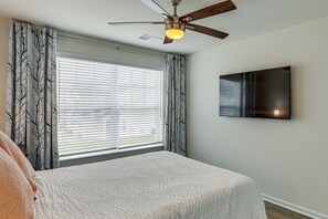 Une chambre à coucher comprenant un lit, un ventilateur de plafond, une fenêtre avec des stores et un téléviseur à écran plat fixé au mur.