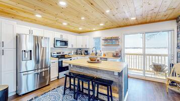A modern kitchen with a wooden ceiling, stainless steel appliances, a large island with stools, and a view to the outside.