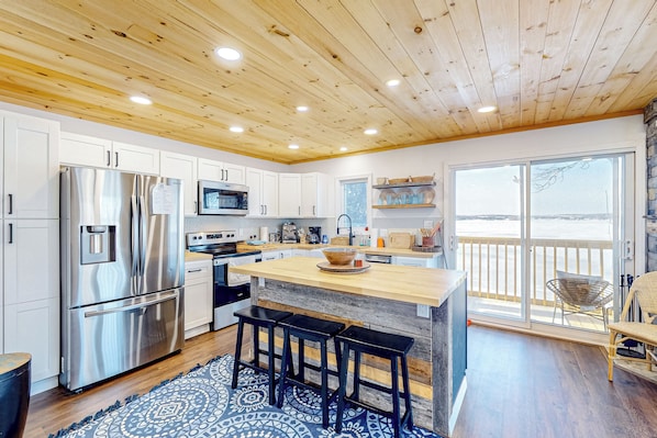 A modern kitchen with a wooden ceiling, stainless steel appliances, a large island with stools, and a view to the outside.