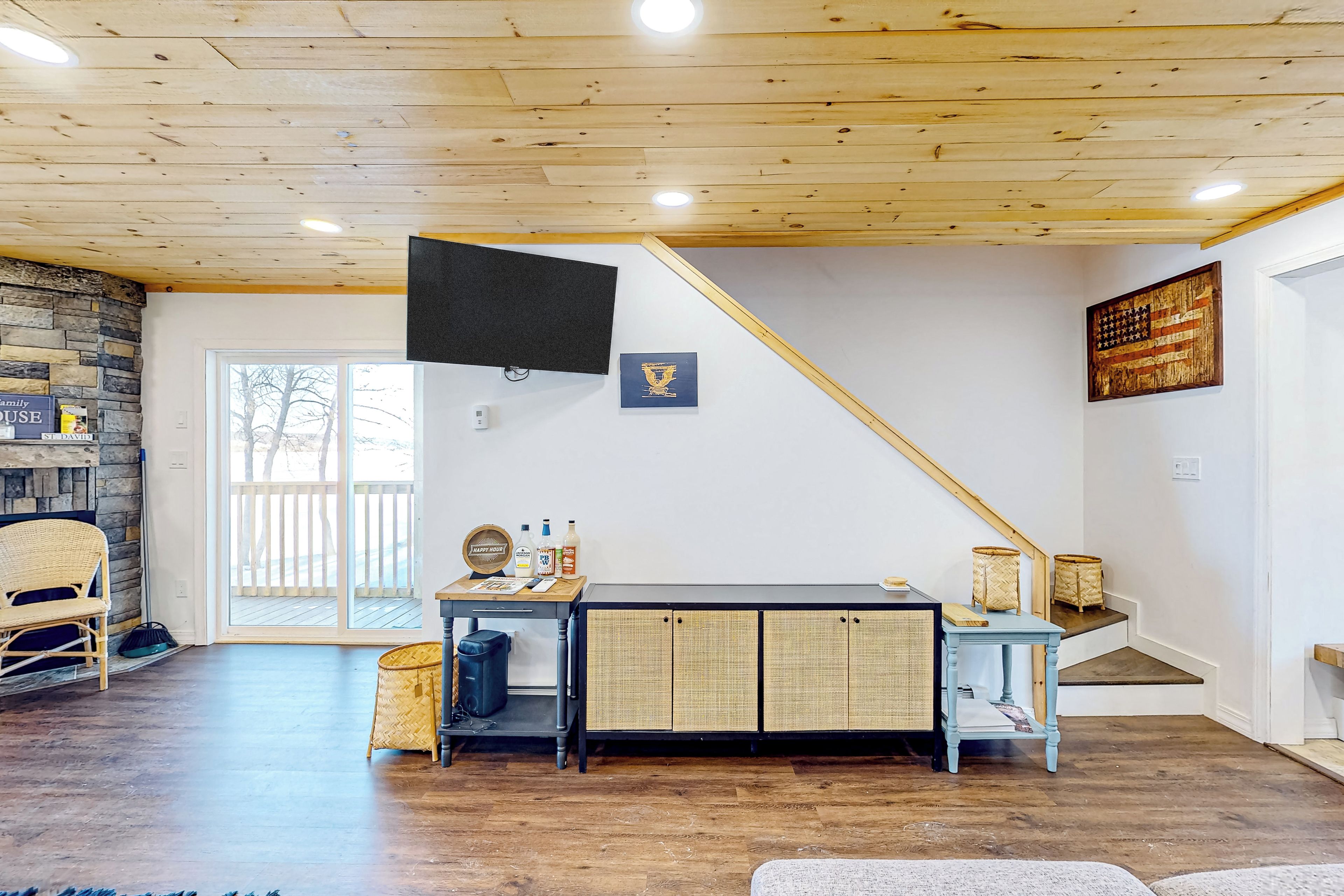 A spacious living room with a wooden ceiling, a stone wall, a large TV, a dining table with wicker baskets, and a staircase.