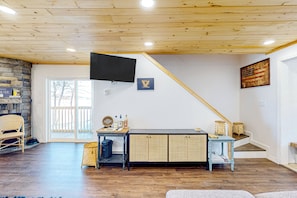 A spacious living room with a wooden ceiling, a stone wall, a large TV, a dining table with wicker baskets, and a staircase. - Waterfront Escape on Long Lake! Ski, Paddle + Play (Saint David)