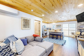 A modern living room with a gray sectional sofa, a wooden ceiling, and a kitchen area with a large window.