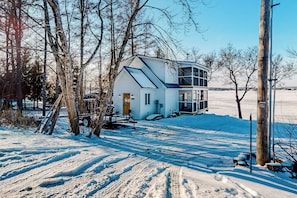 A snow-covered landscape with a two-story house featuring a large window and a balcony. - Waterfront Escape on Long Lake! Ski, Paddle + Play (Saint David)