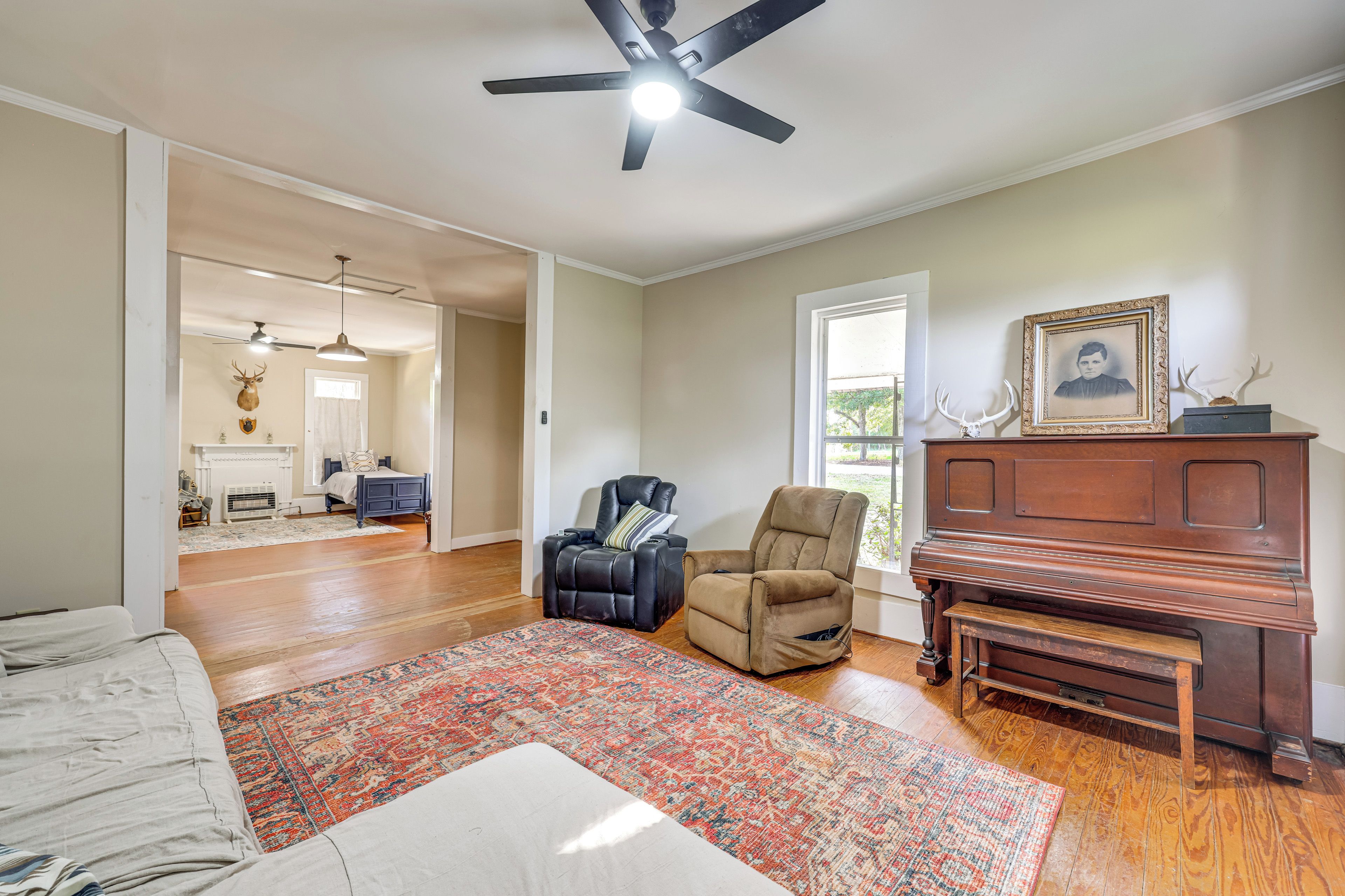A spacious living room with hardwood flooring, a large window, a ceiling fan, a fireplace, and a piano.