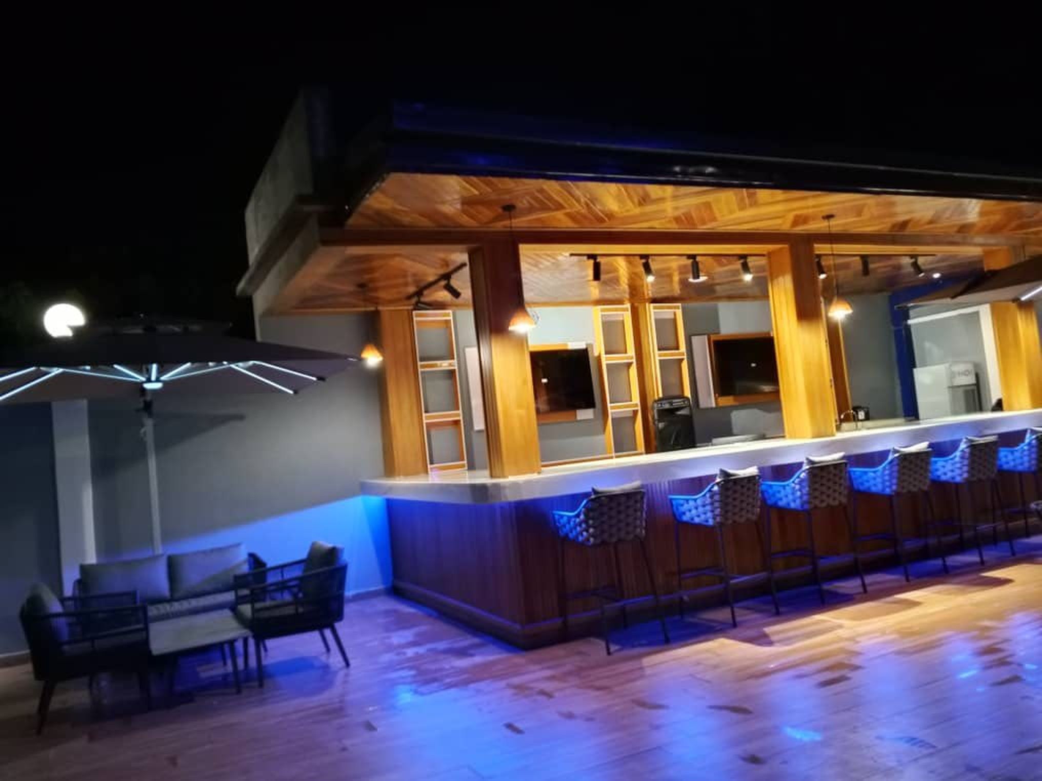 Un área de bar al aire libre con mostrador de madera, taburetes y una iluminación ambiental.