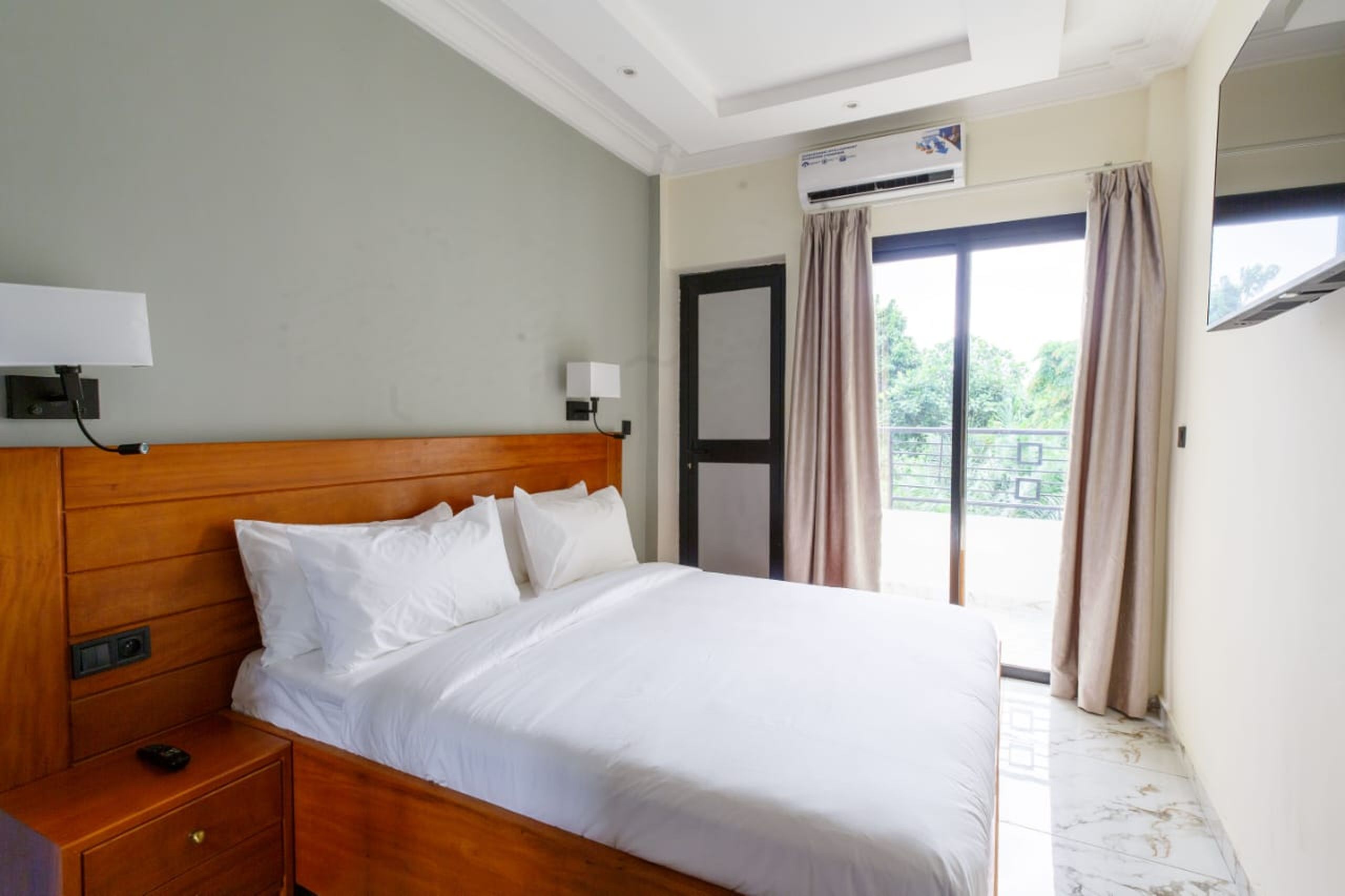 A hotel room with a wooden headboard, a bed with white linens, a nightstand, a wall-mounted lamp, an air conditioning unit, and a window with curtains.