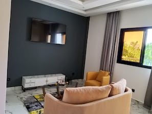 Una sala moderna con un sofá, una mesa de centro, un televisor montado en la pared y un ventanal con vistas a zonas verdes.